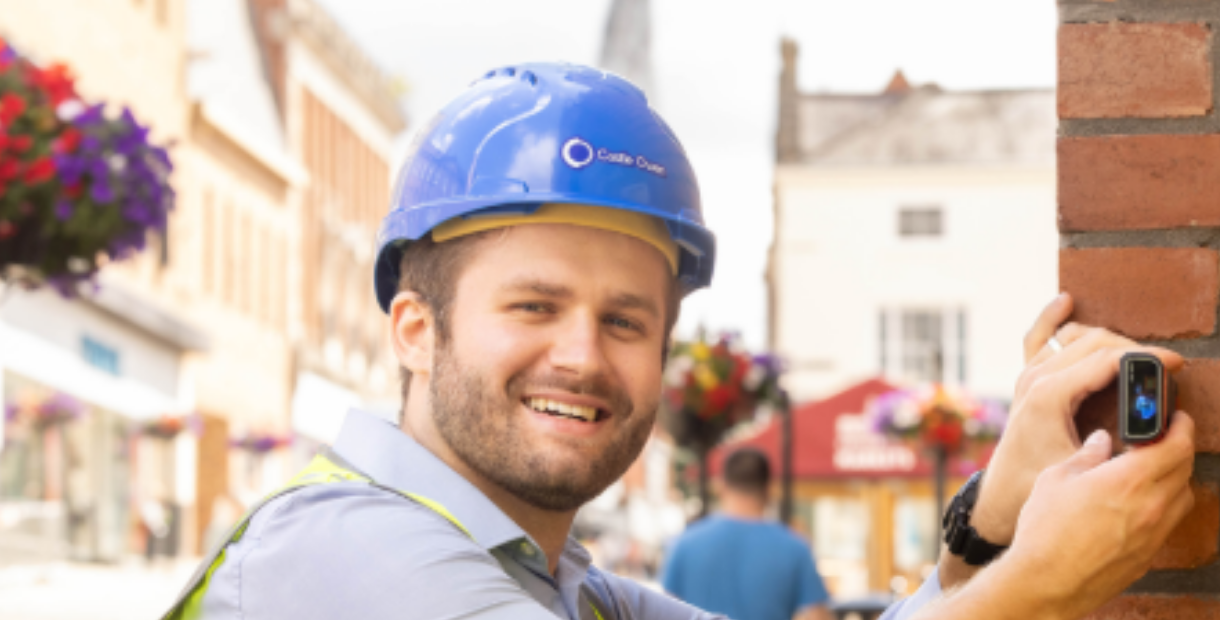 Young man wearing a blue hard hat holding a measuring device against a wall outside