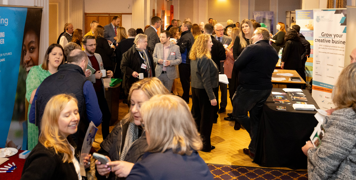 A busy networking event in a large indoor venue, with dozens of attendees standing in small groups and talking around exhibition stands and information tables. Promotional banners and leaflets are displayed throughout the room, including a banner promoting support for creative businesses. The space is brightly lit, with people holding drinks and informational materials while networking.
